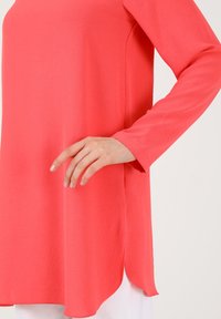 Top in corallo a maniche lunghe con una texture liscia, caratterizzato da un orlo arrotondato e fessure laterali per facilitare i movimenti. Design semplice senza motivi.