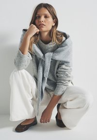 Maglione grigio a maglia con scialle abbinato drappeggiato sulle spalle, abbinato a pantaloni bianchi a gamba larga e scarpe basse marrone scuro.