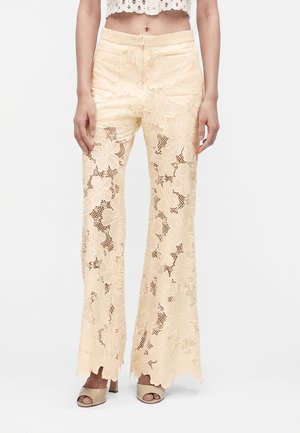 Pantalon en dentelle florale beige à larges jambes et ourlets festonnés, porté avec des sandales à talons ouvertes beige et un top court blanc.