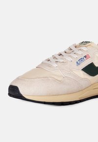Sneaker beige con una combinazione di materiali in suede e mesh, con dettagli verdi, lacci bianchi e una suola in gomma nera. Logo del marchio sul lato.
