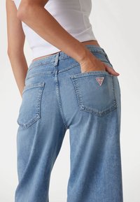 Jeans in denim a gamba larga di colore azzurro chiaro con due tasche posteriori. Presentano una toppa triangolare del marchio sulla tasca destra e dettagli di cucitura in vista.