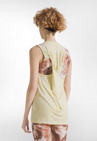 Gul ærmeløs top med draperet ryg, lagdelt over en mønstret brun og pastelfarvet tanktop. Tekstureret stof, afslappet pasform, synlige syningsdetaljer.