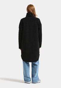 Svart fleece lång jacka med hög krage och sidofickor, med en strukturerad, mjuk yta, bärs över ljusblå vida jeans.