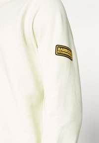 Krémová mikina s měkkou texturou a žebrovanými manžetami, s černou a žlutou nášivkou "Barbour International" na rukávu.