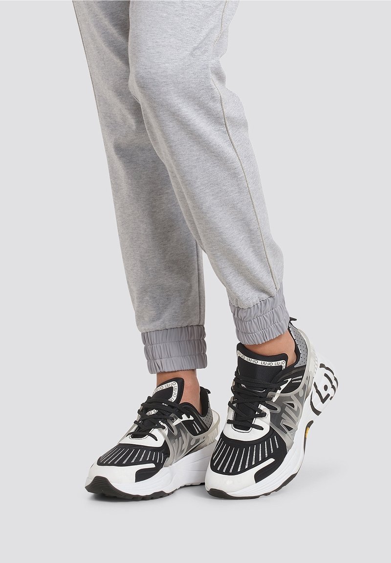 Personne portant un pantalon de jogging gris et des baskets noires et blanches avec des semelles épaisses et des détails à motifs, debout sur un fond uni clair.