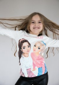 T-shirt bianco a maniche lunghe con illustrazioni di cartoni animati che raffigurano due ragazze con dettagli precisi. I capelli delle ragazze sono mossi e lo sfondo è neutro.