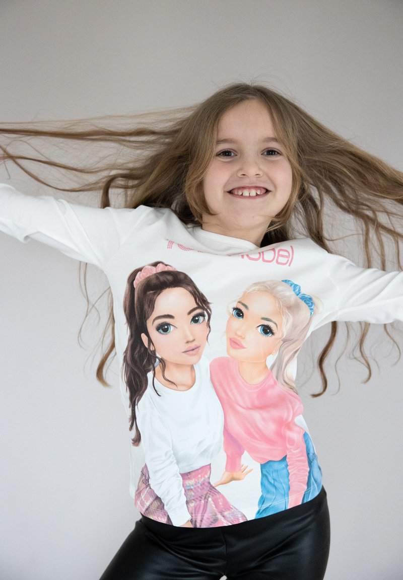 T-shirt bianco a maniche lunghe con illustrazioni di cartoni animati che raffigurano due ragazze con dettagli precisi. I capelli delle ragazze sono mossi e lo sfondo è neutro.
