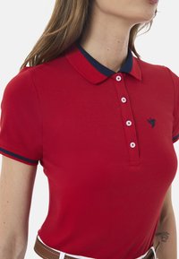 Rød polo skjorte med en marineblå krave, med to-knaps lukning og lille broderet fuglelogo. Korte ærmer med marineblå detaljer.