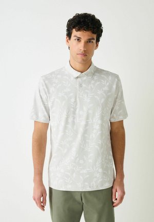 Tânăr cu păr scurt și creț, purtând un tricou polo gri deschis cu model floral și pantaloni verde măsliniu, stând în picioare pe un fundal alb simplu.