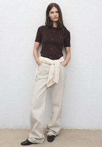 Maglione corto marrone a maniche corte con decorazioni sottili, abbinato a pantaloni beige a gamba larga e un maglione chiaro legato in vita.