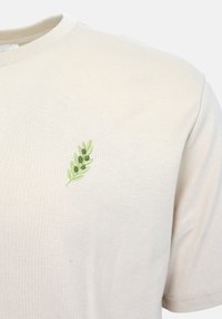 Μπεζ μπλούζα με στρογγυλή λαιμόκοψη και μικρό πράσινο κεντημένο κλαδί ελιάς στο αριστερό στήθος, εμφανίζεται στο πάνω μέρος του κορμού.