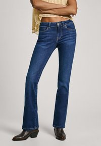 Jeans de mezclilla oscuros de corte acampanado con cintura ajustada, diseño de cinco bolsillos y detalles de costura sutiles, combinados con botines de tacón marrones.