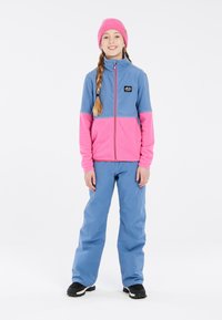 Fleecejack in blauw en roze met een rits aan de voorkant en logo, gecombineerd met blauwe skibroek en zwarte laarzen, op een simpele witte achtergrond.