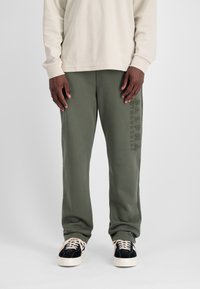 Groene sweatpants met een zachte textuur, voorzien van een verticale reliëflogo op de linker bovenbeen, gecombineerd met zwart-witte sneakers.