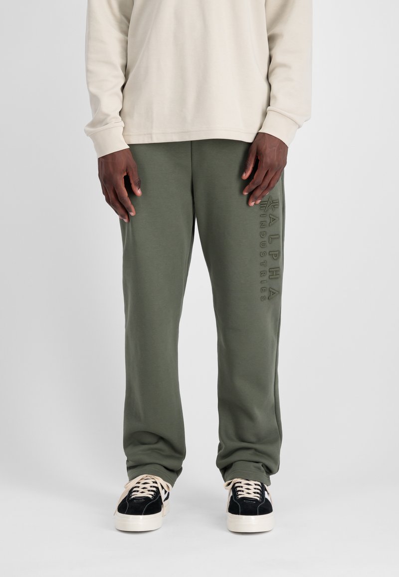 Groene sweatpants met een zachte textuur, voorzien van een verticale reliëflogo op de linker bovenbeen, gecombineerd met zwart-witte sneakers.