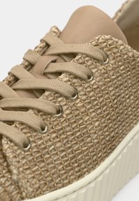Gros plan d’une basket beige avec une texture tissée, des lacets en tissu passés à travers des œillets métalliques et une semelle épaisse en caoutchouc blanc.