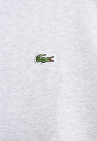 Tejido de sudadera gris con textura suave, que presenta un pequeño logo de cocodrilo verde con detalles en rojo bordado en el lado izquierdo del pecho.