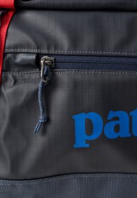 Siva platnena nahrbtnik s modrim logotipom, rdečimi trakovi in zapirnim žepom z kovinsko opremo in teksturirano površino.
