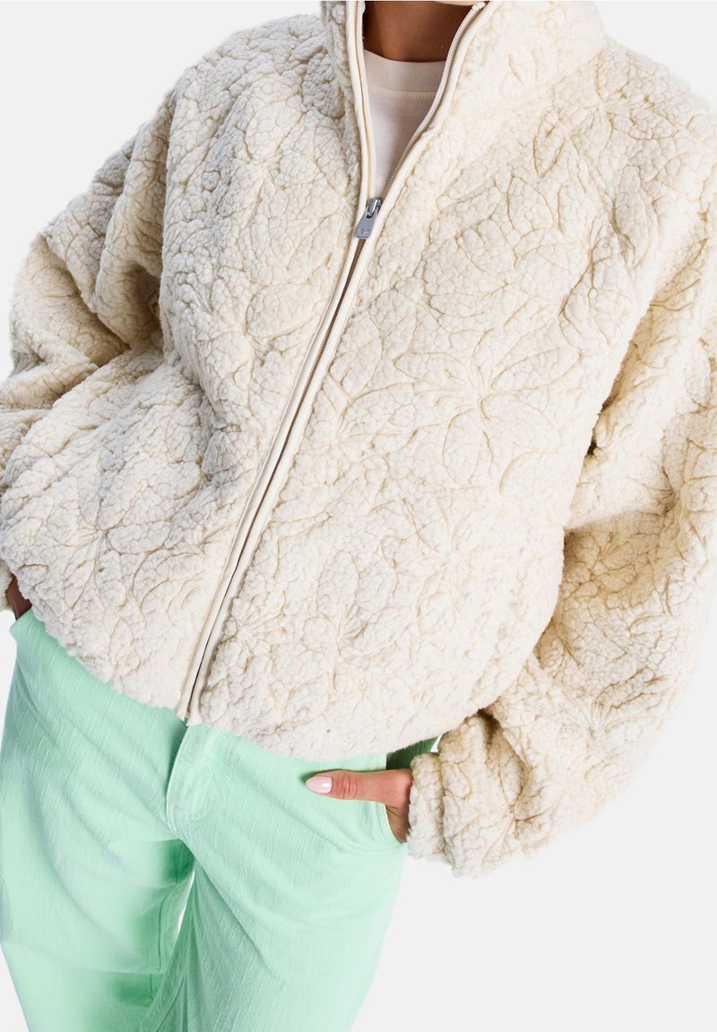 Kremowa, teksturowana kurtka z polaru z zamkiem błyskawicznym i wzorami liści. Charakteryzuje się oversize’owymi rękawami i wysokim kołnierzem, zestawiona z miętowozielonymi spodniami.