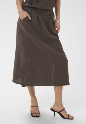 Kobieta nosząca brązową teksturowaną spódnicę midi z elastycznym paskiem i czarne sandały na wysokim obcasie, stojąca z jedną ręką w kieszeni.