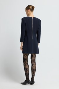 Abito navy con spalle strutturate e zipper nascosto sul retro, abbinato a collant neri trasparenti con motivo floreale e tacchi neri.