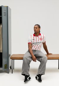 Camisola desportiva branca com riscas vermelhas, logótipo e letras pretas, combinada com jeans cinzentos largos e sapatos pretos com detalhes brancos.