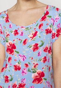 Donna che indossa una camicia azzurro chiaro con un vivace motivo floreale rosso, rosa e verde e scollo a barca.