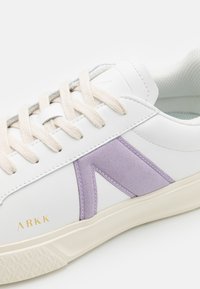 Zapatilla de cuero blanca con un acento de ante lavanda, cordones crema y una suela de goma beige con agarre texturizado. Logo de ARKK en dorado.