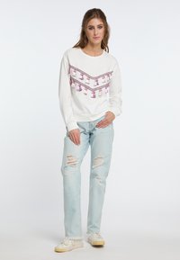 Felpa bianca con decorazioni a frange viola in un motivo a zig zag, abbinata a jeans azzurri strappati e sneakers bianche.