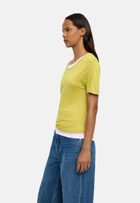 Limonkowa koszulka z krótkim rękawem z różowym ściągaczem, zestawiona z luźnymi niebieskimi dżinsami. Lekki materiał o gładkiej fakturze.