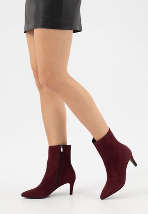 Žena v černé kožené mini sukni a vínových semišových kotníkových botách se zipem na boku a nízkými podpatky na jednobarevném pozadí.