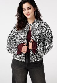 Zwart-wit zebra gestreepte oversized jas met een strik aan de hals, voorzien van volumineuze mouwen en bordeauxrode kantaccenten bij de manchetten.