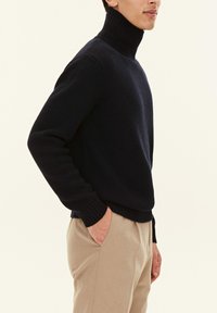 Maglione a collo alto blu navy realizzato in tessuto a maglia, con polsini e orlo a costine, indossato con pantaloni beige chiaro.