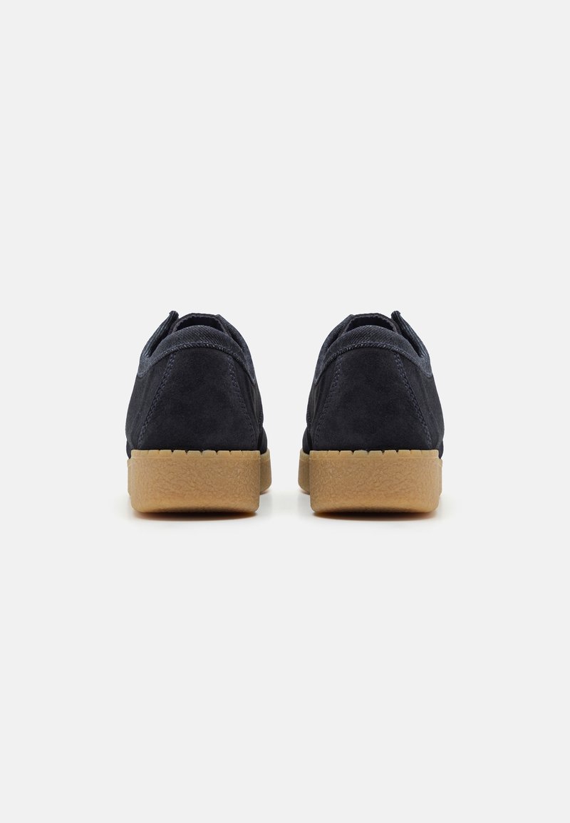 Zapatos de ante azul marino con forma redondeada, que cuentan con una suela de goma y textura suave. Detalle del tacón visible, diseño minimalista.
