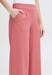 Pantaloni a gamba larga in un morbido tessuto rosa, con vita regolabile tramite cordoncino, tasche laterali e pieghe vicino alla vita per un tocco di dettaglio.