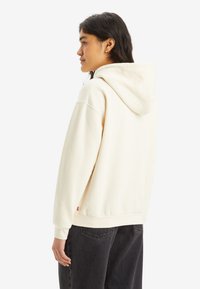 Kvinna med långt mörkt hår klädd i en off-white hoodie och svarta jeans, stående i profil mot en enkel bakgrund.