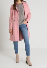 Lång rosa kappa med mjuk textur, silverknappar och knäppta manschetter, i kombination med en grå ribbad tröja och blå jeans. Beigefärgade anklarstövlar.