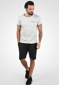 T-shirt gris à manches courtes avec une poche, bord noir et shorts noirs avec cordon de serrage. Porté avec des baskets noires. Texture du tissu lisse.