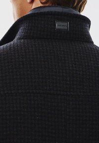 Płaszcz z wełny w ciemnogranatowym kolorze w wzór houndstooth, z wysokim kołnierzem i czarną metką przyszytą z tyłu. Gładka tekstura.