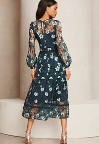 Marineblauwe jurk met bloemenborduursel, transparante lange mouwen, een keyhole rug en een gelaagde rok, met groene bladeren en lichtblauwe bloemen.