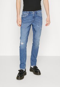 Bleka blå jeans med slitna detaljer, slim fit, tillverkade av denim, med fem fickor och knappstängning. Bärs med svarta skor.