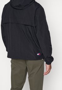 Czarna lekka kurtka z kapturem, posiadająca tylną wstawkę. Wyposażona w elastyczne mankiety oraz naszywkę z logo na dole. Gładka tekstura.