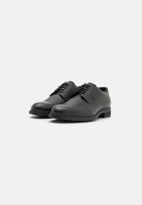 Chaussures de mariage en cuir noir avec une surface texturée, un bout arrondi et un design à lacets. Présentent un petit talon carré et une esthétique minimaliste.