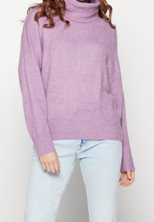Vrouw in een lila coltrui en lichtblauwe spijkerbroek, staande tegen een eenvoudige witte achtergrond.
