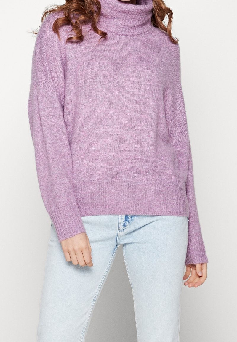 Frau in einem lilafarbenen Rollkragenpullover und hellen Jeans, die vor einem schlichten weißen Hintergrund steht.
