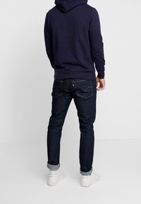 Granatowy hoodie z kieszenią typu kangur, połączony z ciemnoniebieskimi dżinsami i białymi sneakersami. Dżinsy mają delikatne podszycie przy kostce.