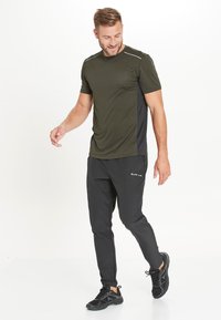 Olivgrünes Sport-T-Shirt mit schwarzen Seiten, Rundhalsausschnitt und reflektierenden Akzenten. Dazu schwarze schmal geschnittene Hose und schwarze Sneakers.