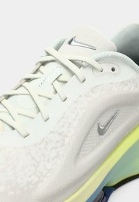 Gros plan sur une basket Nike Vapormax blanche avec une tige en mesh, des lacets blancs et des logos Nike swoosh argentés visibles sur la langue et sur le côté.
