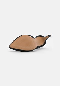 Chaussure à talon haut noire avec bout pointu, semelle beige et talon aiguille fin, posée sur le côté sur fond blanc.