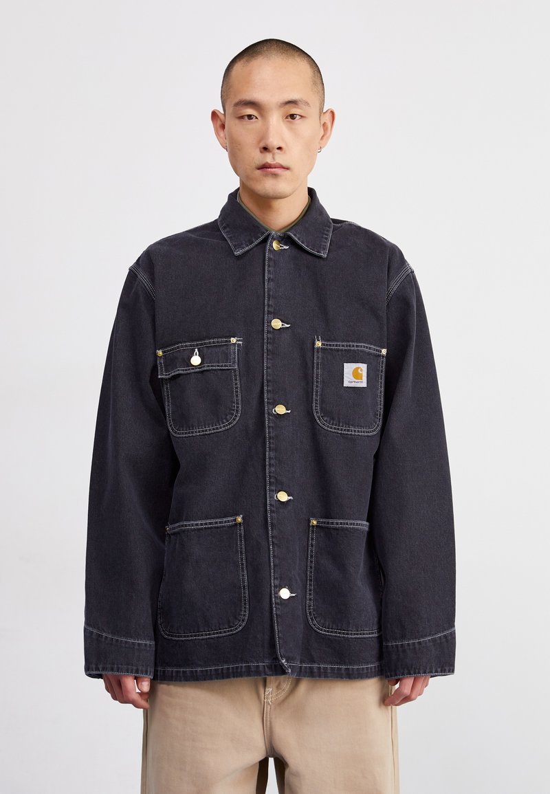 Dark denim jacket with a collared neckline, two front pockets, and gold hardware buttons. Features white stitching and a logo patch.